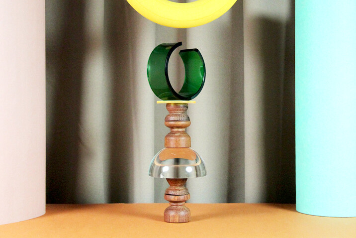 Skulptur ähnlich einer Schachfigur in silberbraun mit gelbem und grünem Ring als KOpf vor einer beigen Vorhangwand