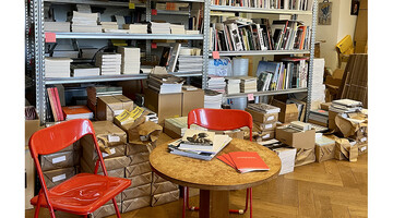 Büroregal mit Büchern, davor ein runder Tisch und zwei orange Plastikklappsessel