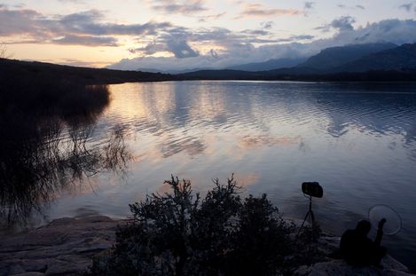 Lake with reflective water surface at sunset, bushes in the foreground and a person with a camera on a tripod at the shore.