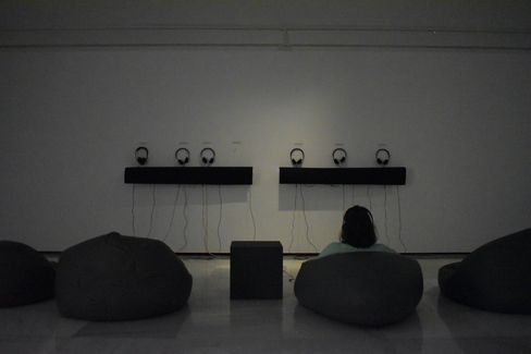 Dimly lit exhibition space with floor cushions and one person seated facing a wall with five headphones mounted.