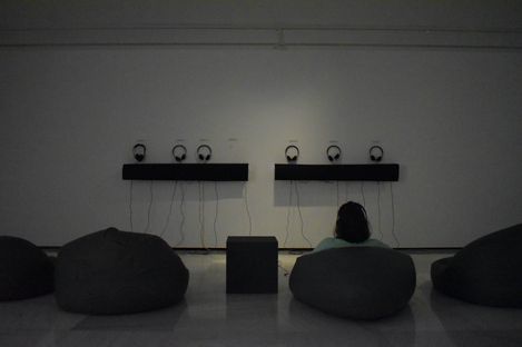 Dimly lit exhibition space with floor cushions and one person seated facing a wall with five headphones mounted.
