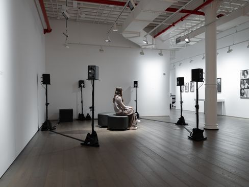 A person sits on a round bench in a white exhibition room surrounded by multiple speakers on stands.