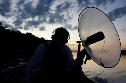 Silhouette of a person wearing headphones sitting by a body of water holding a large round microphone.
