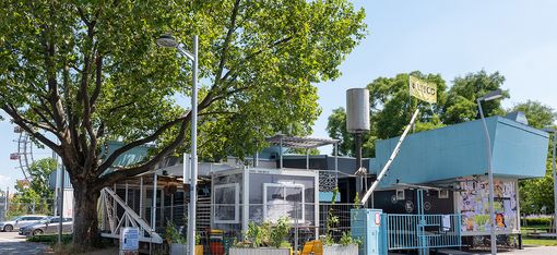 Auf der linken Seite des Bildes steht ein großer Baum, dahinter erahnt man  das Riesenrad. Das Gebäude hat hauptsächlich die Farbe türkis. Es gibt auch viele Glasfronten. Es sieht so aus, als würde sich die Erscheinungsform immer wieder ändern.