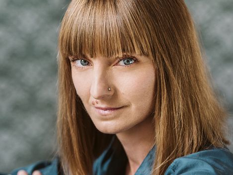 Frau mit schulterlangem, glattem Haar und blauem Hemd sitzt mit verschränkten Armen vor unscharfem Hintergrund.
