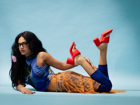 Woman lying sideways on the floor wearing a blue top, an orange mesh dress, and red platform sandals, face blurred.