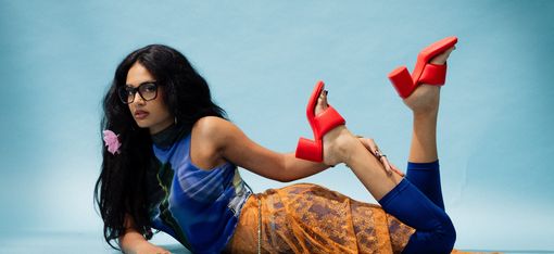 Woman lying sideways on the floor wearing a blue top, an orange mesh dress, and red platform sandals, face blurred.