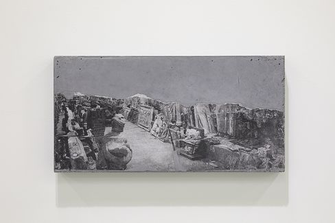 Black-and-white photograph of an ancient ruin with collapsed stone sculptures and walls on a rectangular panel mounted on a white wall.