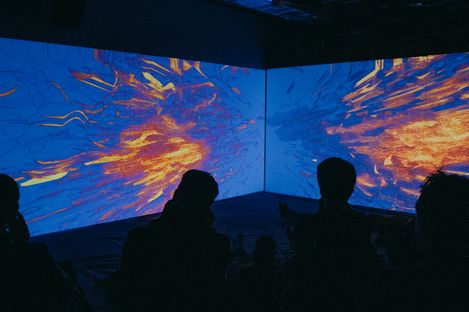 Dark room with two large screens on the walls showing abstract orange and blue patterns with several seated people in silhouette in front.