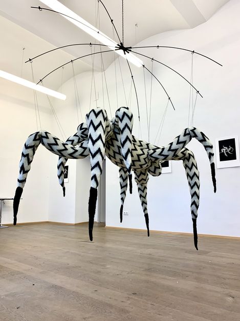 Large hanging art installation with multiple long pointed legs in black-and-white zigzag pattern in a white gallery space.