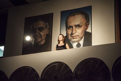 Woman in black sleeveless top standing on a balcony in front of two large portrait paintings on a wall.