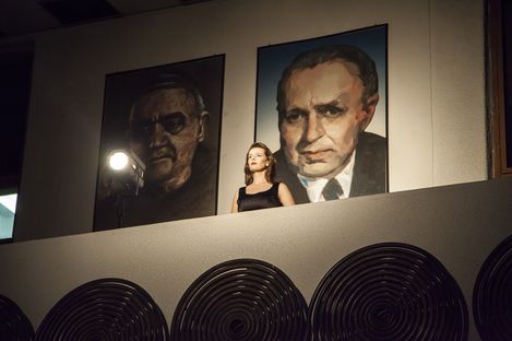 Woman in black sleeveless top standing on a balcony in front of two large portrait paintings on a wall.