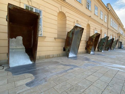 Six large rusty metal funnels leaning diagonally against a building wall with water flowing onto the pavement.