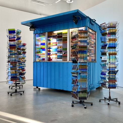 Blue kiosk with large windows, surrounded by several rotating postcard stands with colorful postcards in a bright room