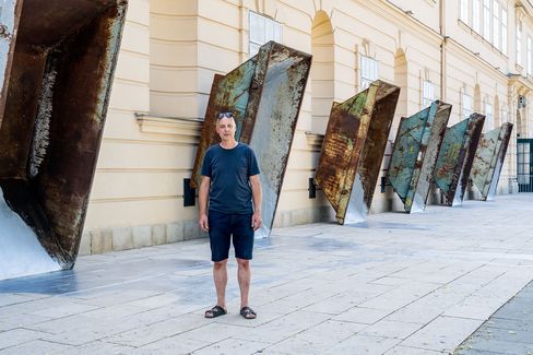 Man in t-shirt, shorts, and sandals standing in front of a row of large rusty metal sculptures attached to a light-colored building facade.
