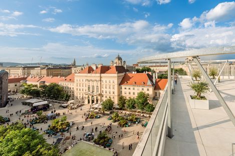 Blick von der Dachterrasse der MQ Libelle auf den belebten Platz des MQ mit historischem Gebäuden und moderner Architektur im Hintergrund