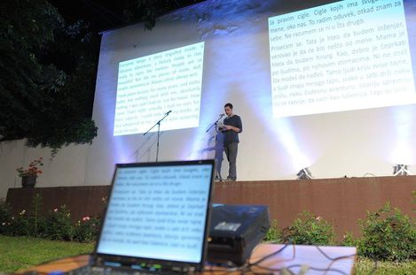 Man standing on stage in front of two large screens projecting text, with a laptop showing text in the foreground out of focus.