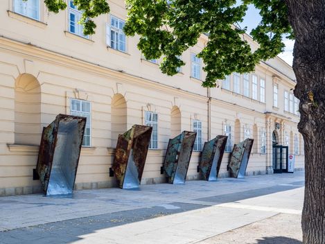 Mehrere große, rostige Metalltrichter lehnen an einer hellen Fassade eines historischen Gebäudes entlang eines Gehwegs.