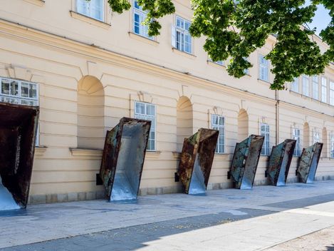 Several large rusty metal funnels leaning against a light-colored facade of a historic building along a sidewalk.