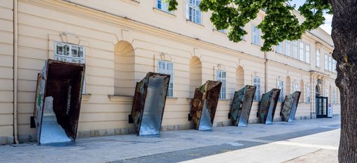 Several large rusty metal funnels leaning against a light-colored facade of a historic building along a sidewalk.