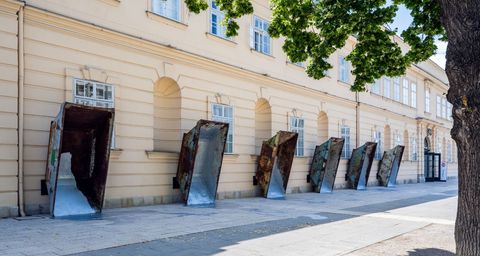 Mehrere große, rostige Metalltrichter lehnen an einer hellen Fassade eines historischen Gebäudes entlang eines Gehwegs.