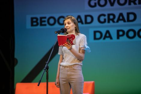 Young woman standing at microphone on stage reading from red book.
