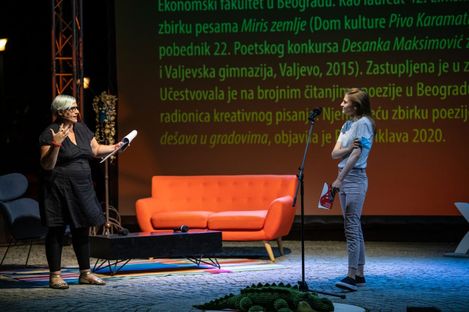 Two people on a stage, one sitting on a chair holding a sheet of paper, the other standing in front of a microphone, with an orange sofa and green text projected on the wall in the background.