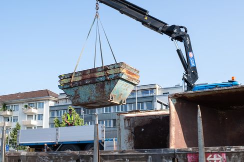 A crane lifts a rusty container over a truck with a worker in high-visibility clothing in front of residential buildings.