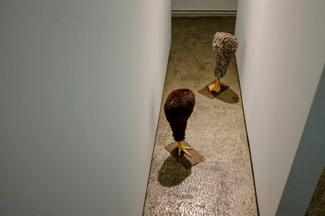 Two headless bird-like sculptures with yellow feet on rectangular bases in a narrow corridor.