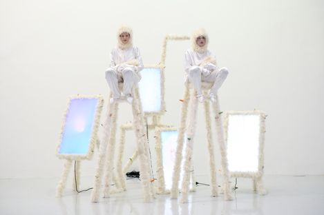 Two people in white protective suits and veils sit on tall ladders wrapped in white material in front of several glowing rectangular screens in a white room.