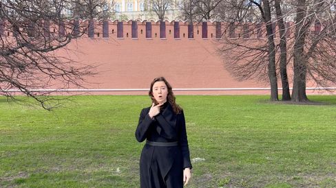 Person wearing black clothes standing on grass in front of a red crenellated wall with trees in the backgroun.