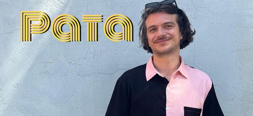 Person with short hair and sunglasses on head wearing a short-sleeved shirt with black and pink halves and a black chest pocket standing against a light wall with the word "Pata".