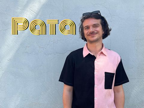 Person with short hair and sunglasses on head wearing a short-sleeved shirt with black and pink halves and a black chest pocket standing against a light wall with the word "Pata".