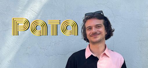 Person with short hair and sunglasses on head wearing a short-sleeved shirt with black and pink halves and a black chest pocket standing against a light wall with the word "Pata".