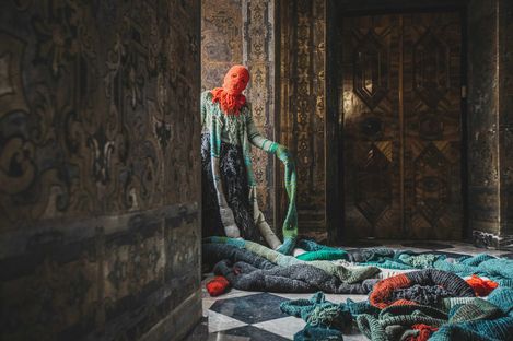 Sculpture of a Medusa with a red head and long twisted snake arms in a historic ornately decorated interior with marble floor and wooden doors.