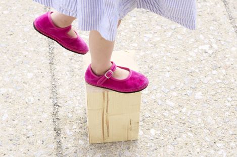 Beine eines Kindes mit schwarzen Sneakers und weißen Socken mit Hundemotiv auf einem gelben Skateboard, das auf einem Holzblock steht