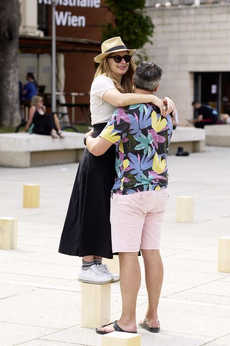 Frau mit Sonnenhut und Sonnenbrille steht auf einem kleinen Sockel und wird von einem Mann in buntem Hemd und Shorts umarmt