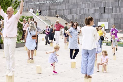Mehrere Menschen unterschiedlichen Alters stehen und sitzen auf einem Platz vor einem modernen Gebäude mit der Aufschrift 'museum moderner kunst stiftung ludwig wien', einige balancieren auf kleinen Holzsockeln
