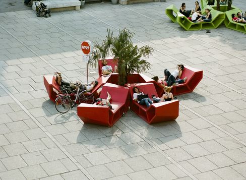 People relaxing on red Enzo seating around a palm tree at MuseumsQuartier Vienna, some lying or sitting back, with a bicycle parked nearby.