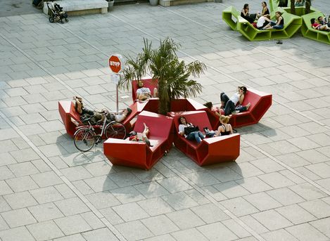 People relaxing on red Enzo seating around a palm tree at MuseumsQuartier Vienna, some lying or sitting back, with a bicycle parked nearby.