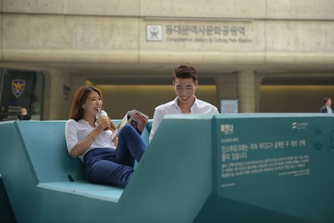 Zwei Personen sitzen entspannt auf einem Enzi-Sitzmöbel und lesen in einem belebten Stadtraum.