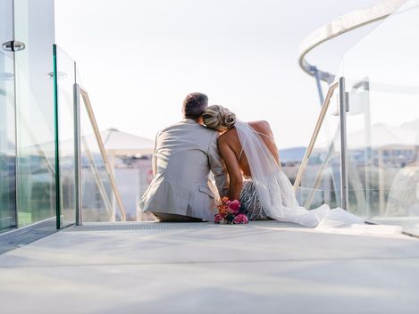 Trauung MQ Libelle, MuseumsQuartier Wien. Ein Ehepaar in Hochzeitskleidung sitzt auf einer Stiege der MQ Libelle und blickt über die Dächer Wiens.