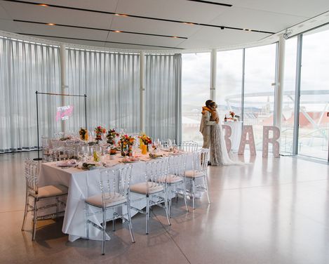 Trauung MQ Libelle, MuseumsQuartier Wien. Braut und Bräutigam genießen einen zärtlichen Moment vor großen Fenstern in einem hellen, modernen Veranstaltungsort. Ein langer Tisch ist elegant mit Blumen und einem auffälligen Barschild gedeckt.