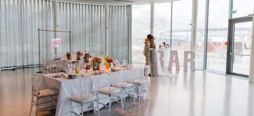 Wedding MQ Libelle, MuseumsQuartier Wien. A bride and groom share a tender moment by large windows in a bright, modern venue. A long table is elegantly set with flowers and a bold bar sign.