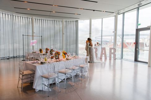 Wedding MQ Libelle, MuseumsQuartier Wien. A bride and groom share a tender moment by large windows in a bright, modern venue. A long table is elegantly set with flowers and a bold bar sign.