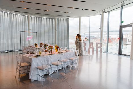 Wedding MQ Libelle, MuseumsQuartier Wien. A bride and groom share a tender moment by large windows in a bright, modern venue. A long table is elegantly set with flowers and a bold bar sign.