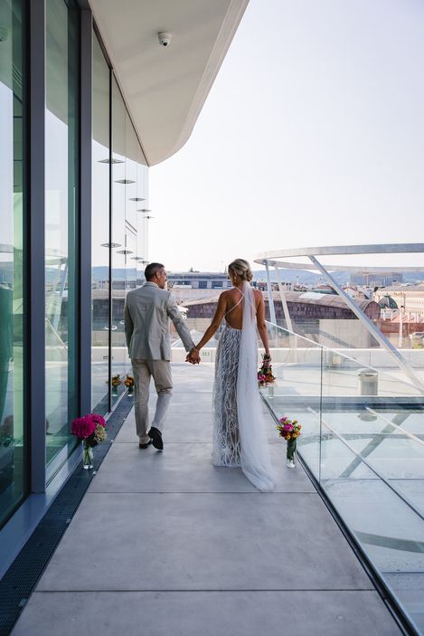 Wedding ceremony at MQ Libelle, MuseumsQuartier Vienna. A married couple walks across the MQ Libelle, which is decorated with flowers. The rooftops of Vienna can be seen in the background.