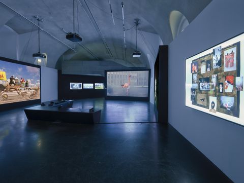 Dimly lit exhibition room with multiple large image projections on the walls and a bench in the center