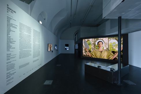 Dark exhibition space with white wall displaying text and multiple screens showing video installations