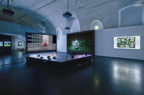Dimly lit exhibition space with multiple large screens showing videos and photos and a platform with headphones in the center